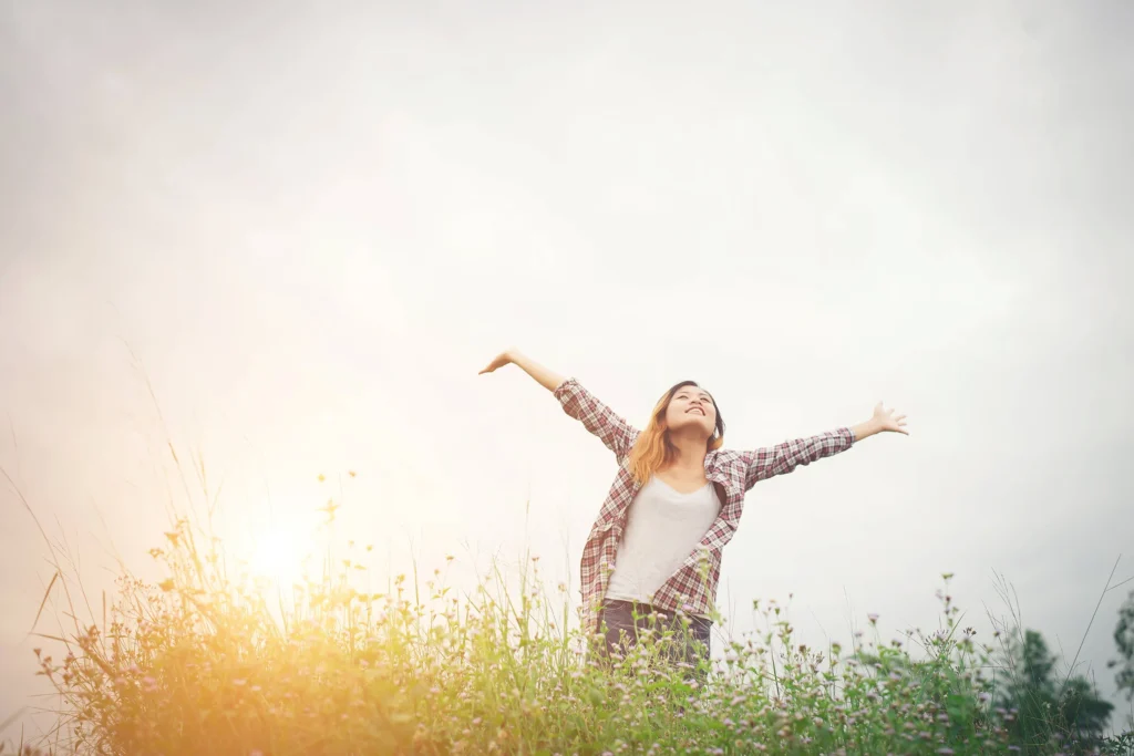 Junge Frau mit ausgebreiteten Armen, steht in einem Blumenfeld bei Sonnenuntergang, symbolisiert Freiheit, Freude und die Verbindung zur Natur.